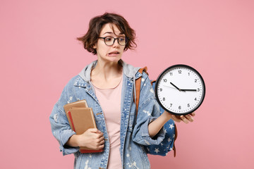 Wall Mural - Confused young woman student in denim clothes eyeglasses, backpack posing isolated on pastel pink background. Education in high school university college concept. Mock up copy space. Hold books clock.