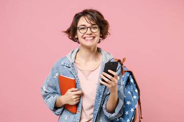 Wall Mural - Smiling young woman student in denim clothes glasses backpack isolated on pastel pink background. Education in high school university college concept. Hold books using mobile phone typing sms message.