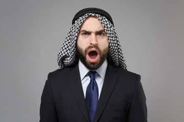 Shocked young bearded arabian muslim businessman in keffiyeh kafiya ring igal agal classic black suit shirt isolated on gray background. Achievement career wealth business concept. Keeping mouth open.