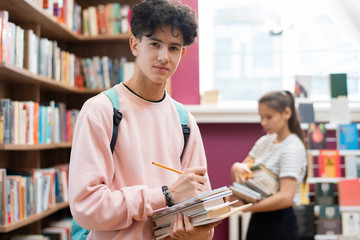 Poster - Clever teenager in sweatshirt looking at you while standing by bookshelf in college library and making notes in notepad