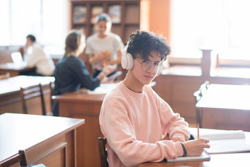 Poster - Youthful clever student of college looking at you through eyeglasses while listening to music in headphones and working by desk