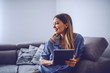 © Dusan Petkovic - Young charming smiling caucasian brunette in stripped shirt sitting on sofa in living room, holding tablet and looking away.