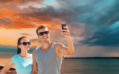 Poster - fitness, sport and healthy lifestyle concept - happy couple in sports clothes and sunglasses taking selfie by smartphone over sea and sunset sky background