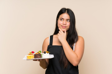 Wall Mural - Young teenager Asian girl holding lots of different mini cakes over isolated background thinking an idea