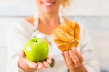 Close Up Of Beautiful Woman Holding An Apple And Croissant In Front Of The Camera Choosing Between A Healthy Life Of Unhealthy Lifestyle - Dieting And Taking Care Of The Food