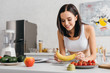 Selective focus of smiling sportswoman putting banana on scales near measuring tape, fruits and notebook on kitchen table, calorie counting diet