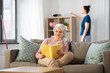 © Syda Productions - leisure, old age and people concept - happy senior woman reading book and housekeeper cleaning at home