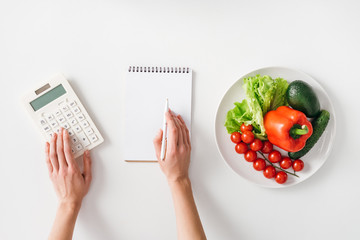 Wall Mural - Top view of woman using calculator near notebook and fresh vegetables on plate on white background