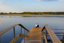 A Romatic Young Couple - The Guy And The Girl Sit Hugged On A Wooden Dock In The Lake. Romantic Meeting.