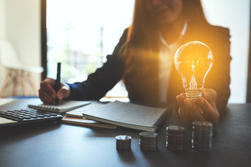 Wall Mural - businesswoman holding a lightbulb with coins stack on table, saving energy and money concept