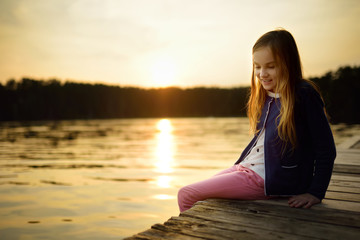 Wall Mural - Cute girl sitting on a wooden platform by the river or lake dipping her feet in the water on warm summer day. Family activities in summer.