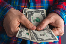 Man In A Red And Blue Plaid Shirt Hands Holding Pile Of Money. Pile Of One And Two Dollars Banknotes In Man Hands