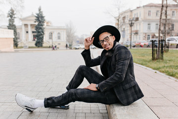 Enthusiastic african guy in white sneakers sitting on the road. Outdoor portrait of black young man in sport shoes chilling on the street.