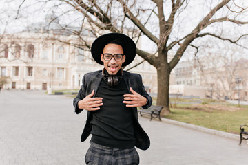 Wall Mural - Smiling african man in checkered pants posing at square. Outdoor photo of glad boy in hat and headpones chilling on street background.