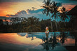 Woman relaxes in a luxury infinity pool overlooking the jungle at sunset in Ubud, Bali. A girl sits on the edge of the infinity pool against the backdrop of a bright beautiful sunset and the jungle.