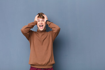 young blonde man raising hands to head, open-mouthed, feeling extremely lucky, surprised, excited and happy isolated against flat wall