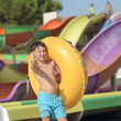 Cute European boy in blue swimming shorts with colorful floater against plastic water slides in Water Park in Spain. He is smiling to the camera and shows like gesture.