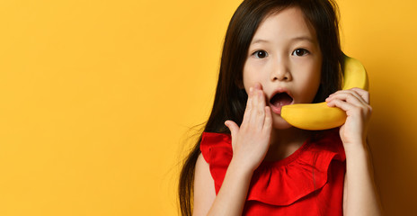 Little asian kid in red blouse. She pretending to be talking on banana like by phone, posing on orange background. Close up