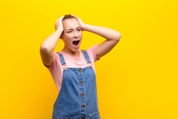 young blonde pretty girl with open mouth, looking horrified and shocked because of a terrible mistake, raising hands to head against yellow wall