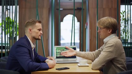 Side view of two middle aged business partners looking at programming code on laptop screen while discussing project details over coffee at cafe table and shaking hands agreeing on deal