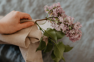 Close-up of hand holding purple lilac flowers branch in bloom on blue linen sheet with neutral linen towel, eco lifestyle concept, still life, vintage effect with grain