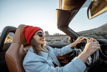 Happy Woman In Red Hat Driving Convertible Car While Traveling On The Desert Road With Beautiful Rocky Landscape On The Background During A Sunset