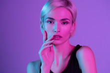 Beautiful  European Girl With Short Blond Hair Wearing Black Top Touching Cheek And Lips With Hand Fingers. Sensual Woman In Neon Uv Light Studio Portrait. Fashion And Art Design Headshot