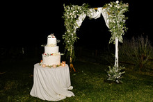 Delicious Creamy Wedding Cake Decorated With Greenery And Flowers On The Table And  Minimal Decorated Archway On The Grass In The Evening
