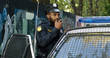 © VAKSMANV - African American young cop in hat and uniform working at patrolling. Policeman standing at his police car and speaking in walkie-talkie. Cop talking in ratio.