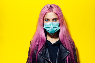 Wall Mural - Studio portrait of teenager girl with pink hair and blue eyes, wearing medical flu mask, dressed in black leather jacket on yellow background.