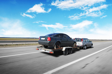 Canvas Print - A crossover with trailer towing a car on the countryside road in motion against sky with clouds