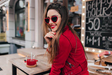 Fascinating girl in heart sunglasses looking over shoulder while sitting at the wooden table. Pleasant brunette woman in red jacket have a good time in street cafe.