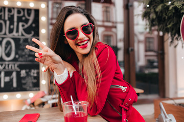 Blissful girl with light-brown hair making peace sign in cafe. Elegant caucasian lady in red jacket relaxing in stylish restaurant with cup of tea.