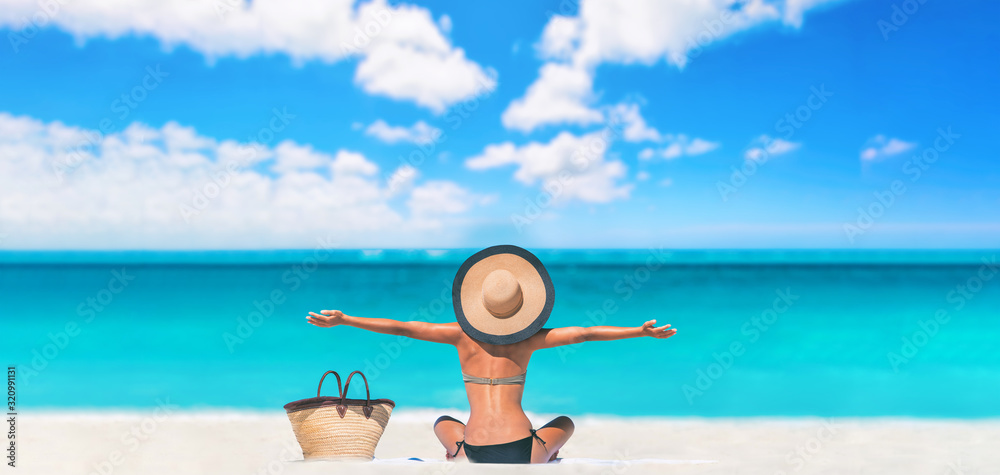 Beach woman happy carefree with open arms on Caribbean travel vacation tourist sun tanning sunbathing on towel body skincare sun exposure protection with hat enjoying summer holidays. - obrazy, fototapety, plakaty 