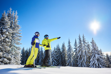 Wall Mural - Low angle shot of skiers resting during skiing in sunny winter weather. Male skier showing sun high in blue sky over winter mountains. Fantastic recreational area of ski resort on background.