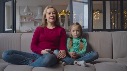 pretty relaxed mother and adorable little girl sitting comfortably cross-legged on sofa and watching