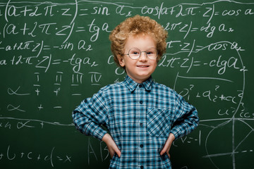 Wall Mural - happy kid in glasses standing with hands on hips near chalkboard