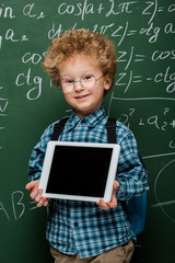 Wall Mural - cheerful kid in glasses holding digital tablet with blank screen near chalkboard with mathematical formulas