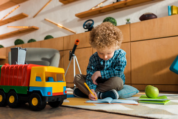 Wall Mural - smart kid writing in notebook while sitting on floor near apple, toy car and books