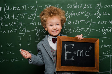 Wall Mural - smiling and smart kid in suit and bow tie holding small blackboard with formula near chalkboard