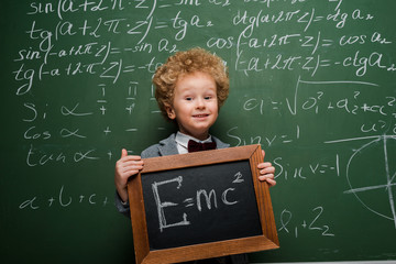 Wall Mural - happy kid in suit and bow tie holding small blackboard with formula near chalkboard