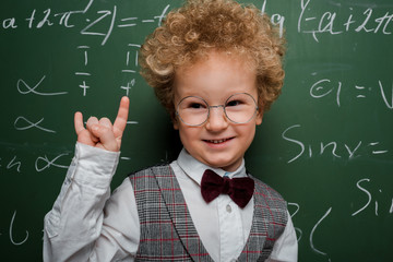 Wall Mural - happy and smart child in suit and bow tie showing rock sign near chalkboard with mathematical formulas