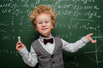 Wall Mural - smiling kid in suit and bow tie holding chalk and pointing with hand at chalkboard with mathematical formulas
