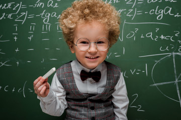 Wall Mural - smart child in suit and bow tie holding chalk near chalkboard with mathematical formulas
