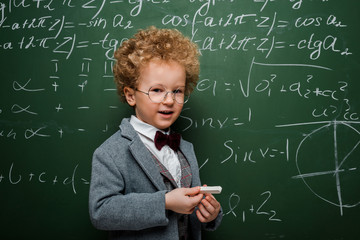 Wall Mural - cute and smart child in suit with bow tie holding chalk near chalkboard with mathematical formulas