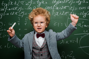 Wall Mural - smart kid in suit with bow tie holding chalk near chalkboard with mathematical formulas