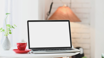 Computer laptop with white blank screen putting together with potted plant and coffee cup on the modern table with comfortable living room as background.