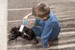 Chłopiec w wieku szkolnym siedzi na dywanie i bawi się z kotem. Kot leży na dywanie zadowolony z pieszczot.