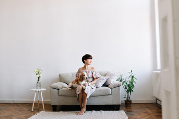 Pretty short-haired lady thoughtfully looking away while beagle dog resting on her knees. Indoor portrait of beautiful girl in pink dress chilling on sofa with cute pet on light background.