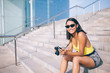 Portrait of happy female photographer in sunglasses sitting on city stairs with modern SLR equipment in hands and smiling at camera, cheerful Latin woman amateur with technology for making content